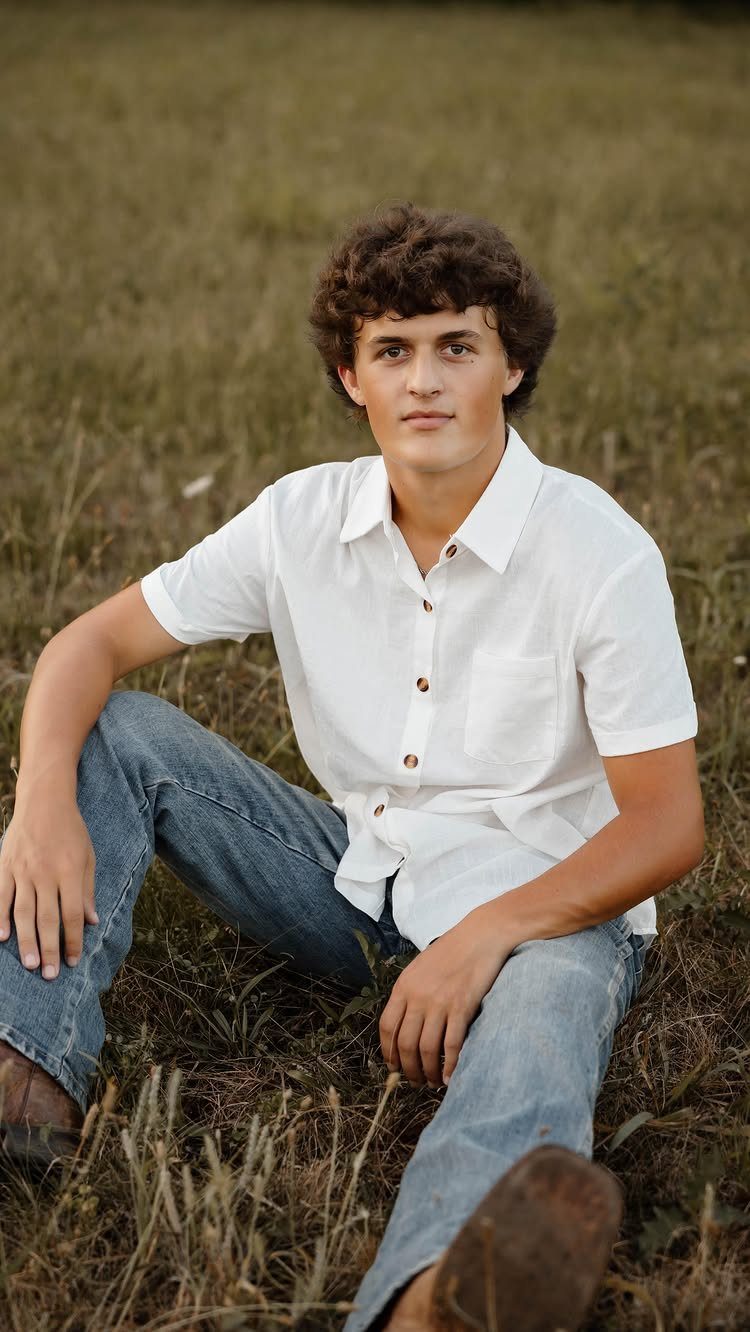 Young man in white shirt and jeans sitting in a grassy field.