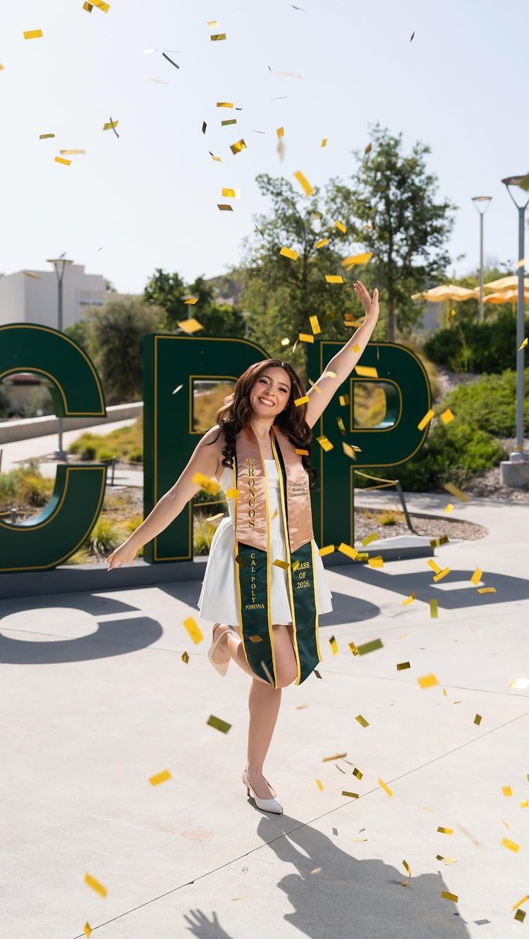 Graduating woman in white dress smiling as yellow confetti falls around her outdoors.