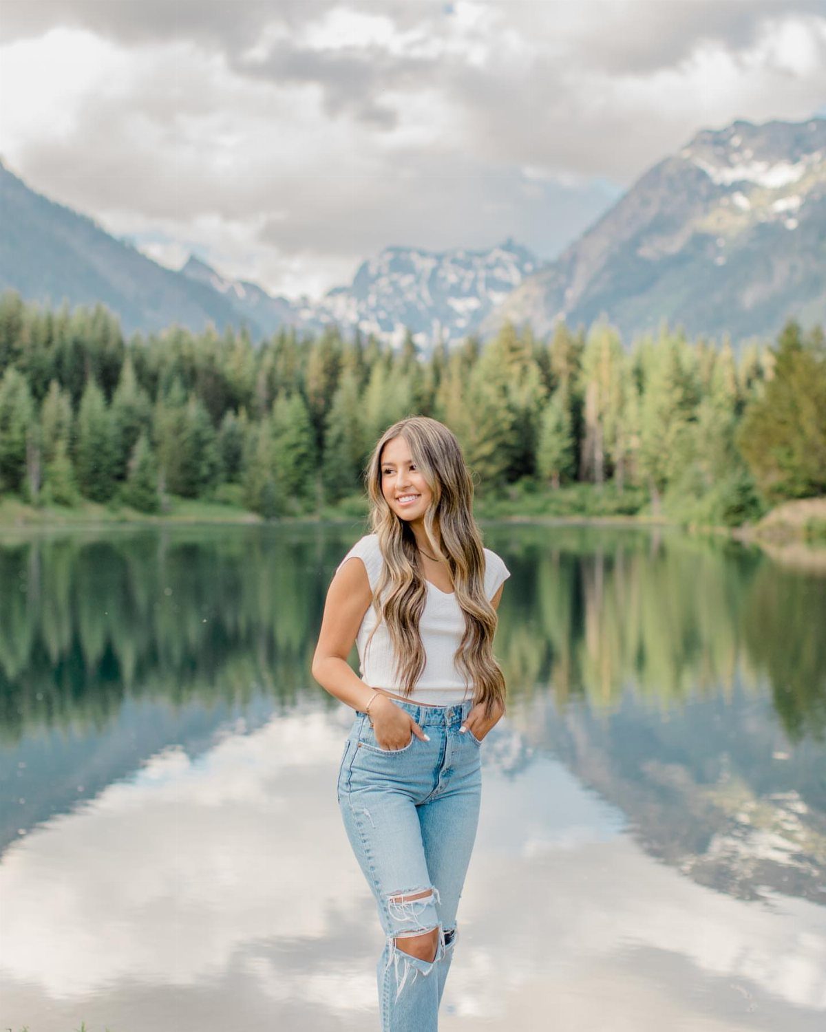 Young woman posing by a reflective lake with mountains and forest in background