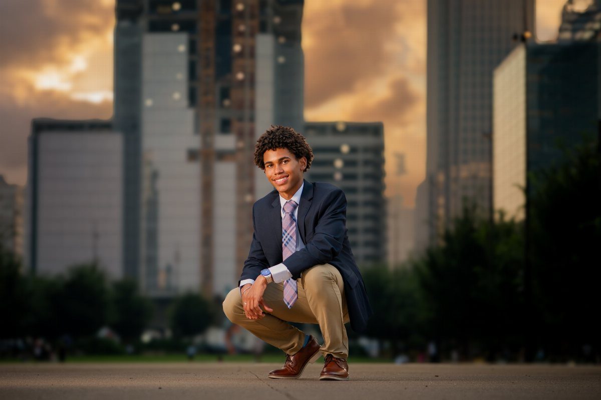 Young man in a blazer crouching in a city plaza with skyscrapers behind him.