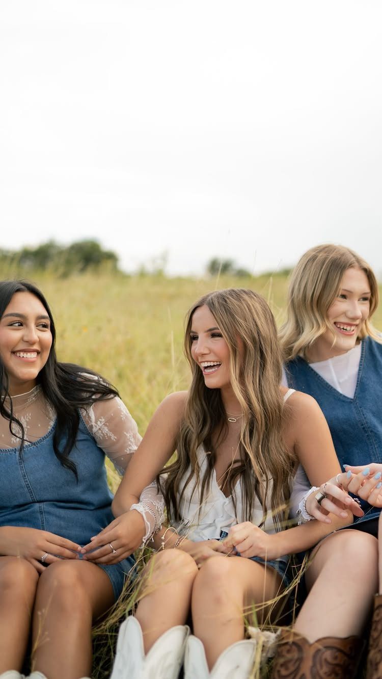 Three smiling young women sitting together in a golden grassy field.