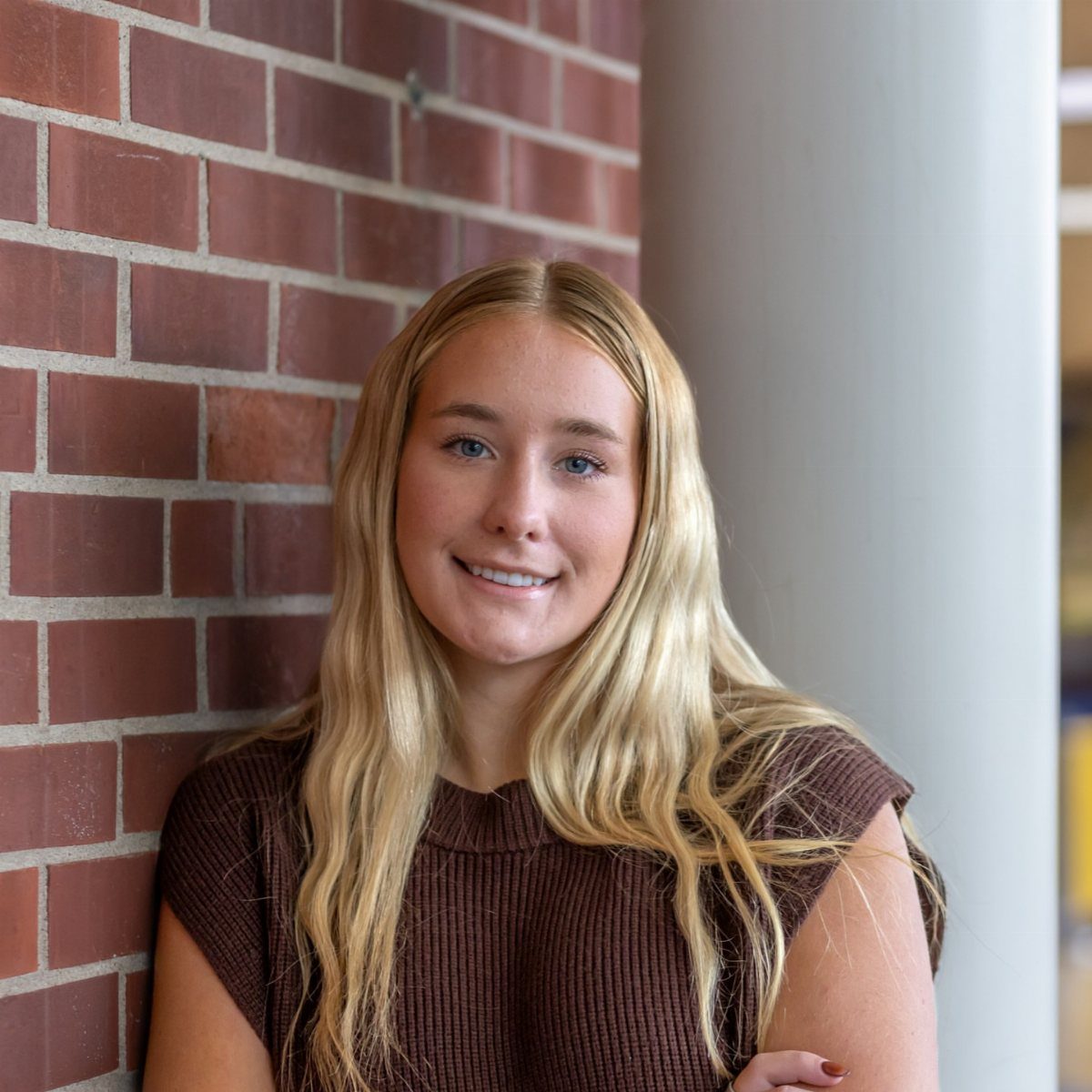 Young blonde woman smiling while leaning against a red brick wall backdrop.