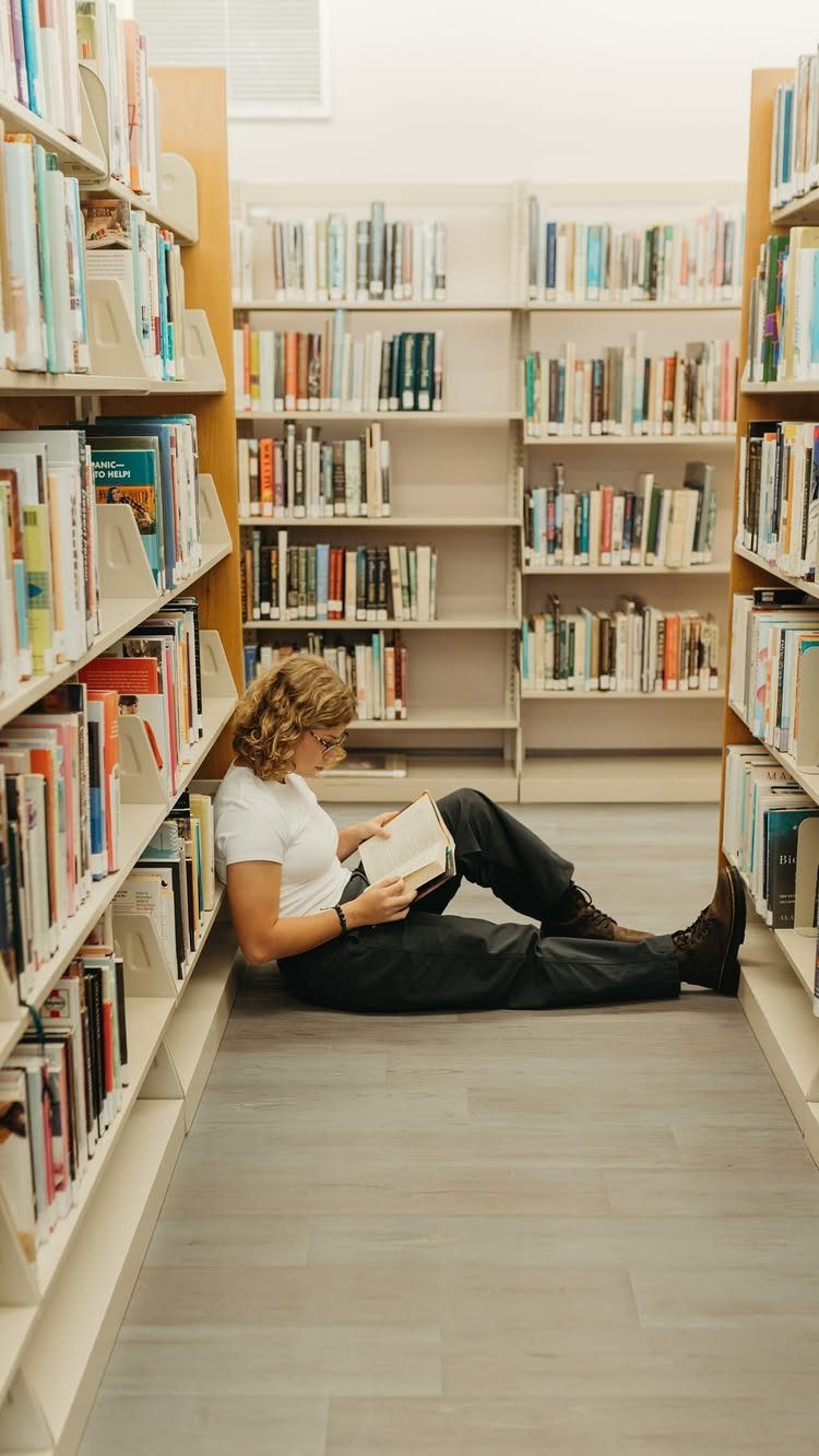 Person sitting on a library floor reading a book between tall bookshelves.