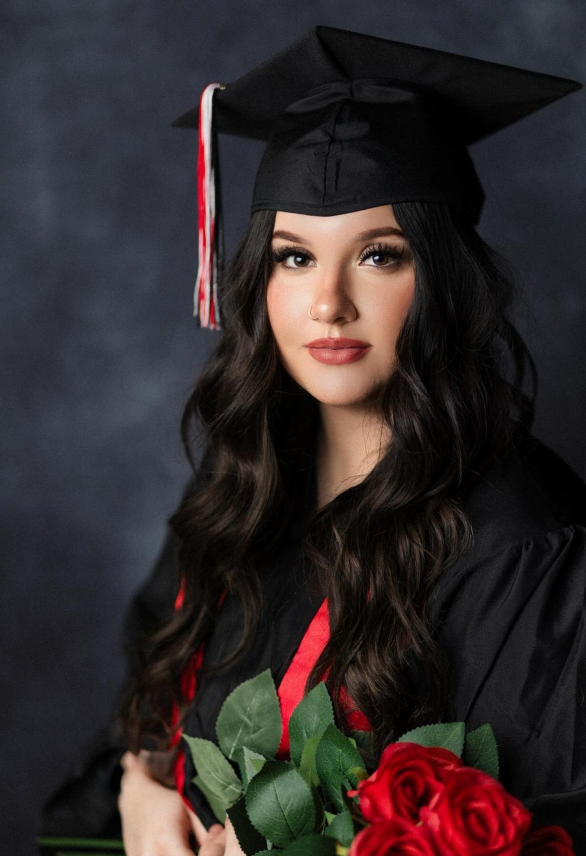 Graduate with wavy dark hair and makeup holding red roses in studio portrait