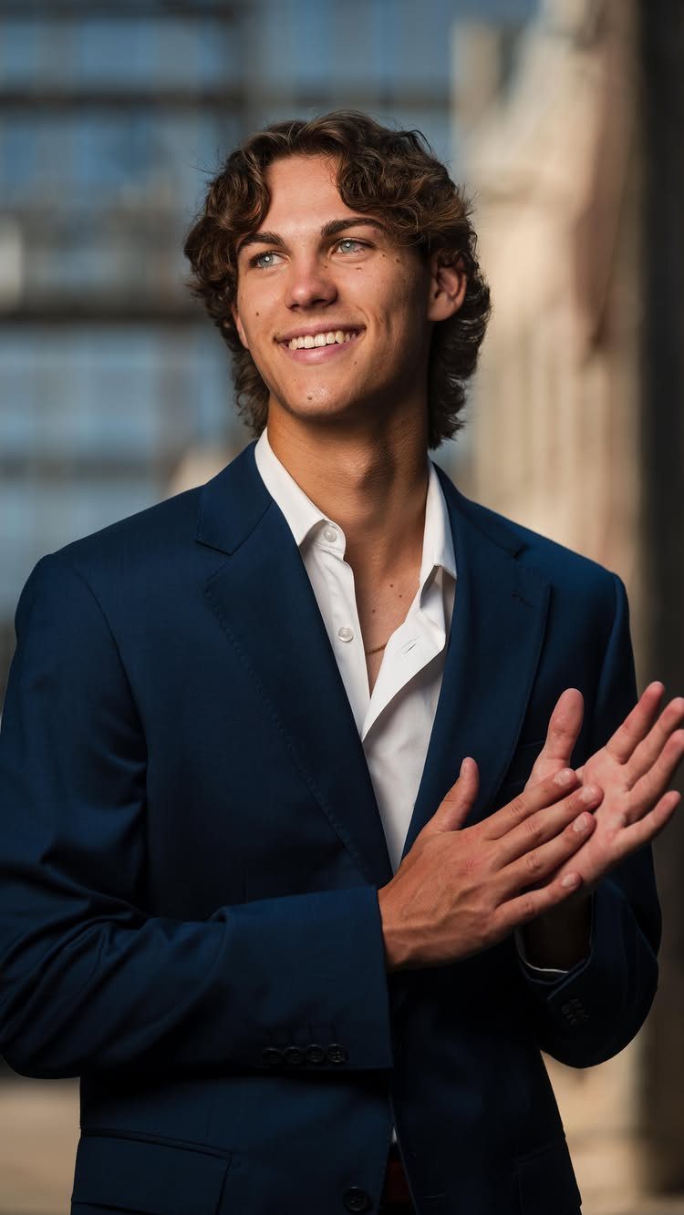 Young man in a navy blue blazer and white shirt smiling outdoors.