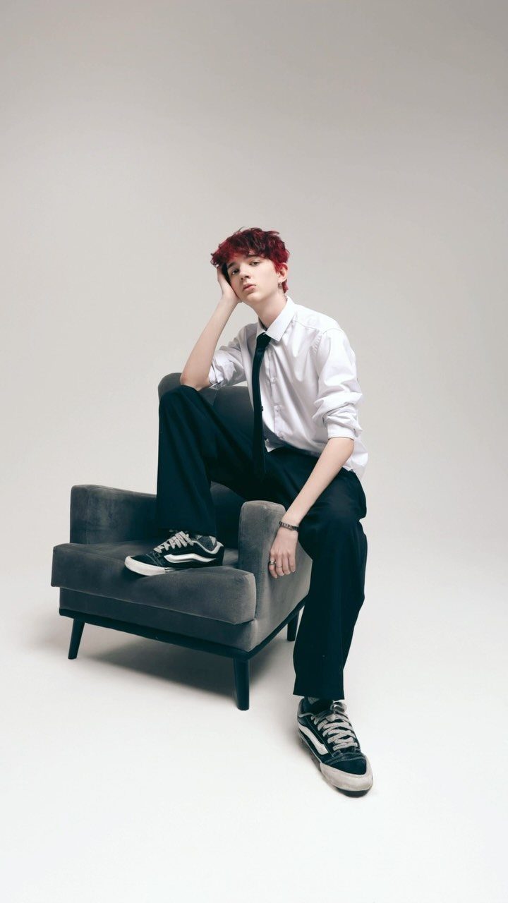 Young person with red hair in formal attire posing with a grey armchair.