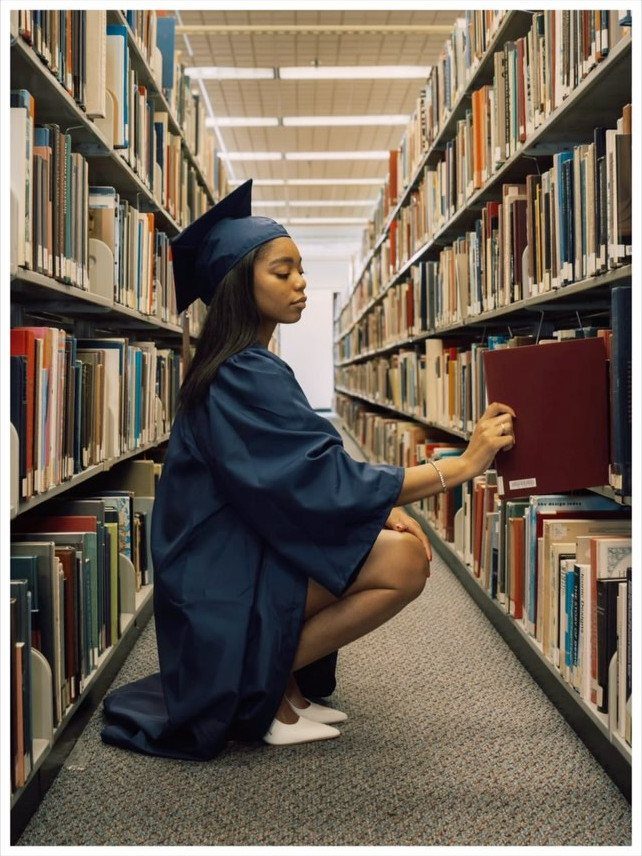 Graduate in navy cap and gown crouching in library aisle reaching for book