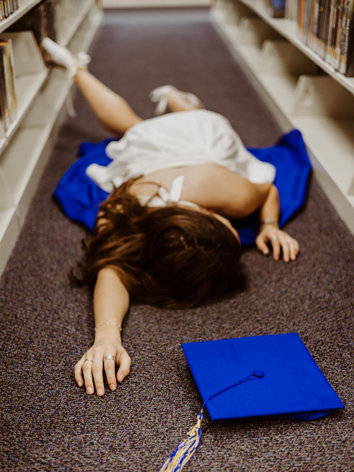Woman lying on library floor in white dress and blue graduation gown and cap