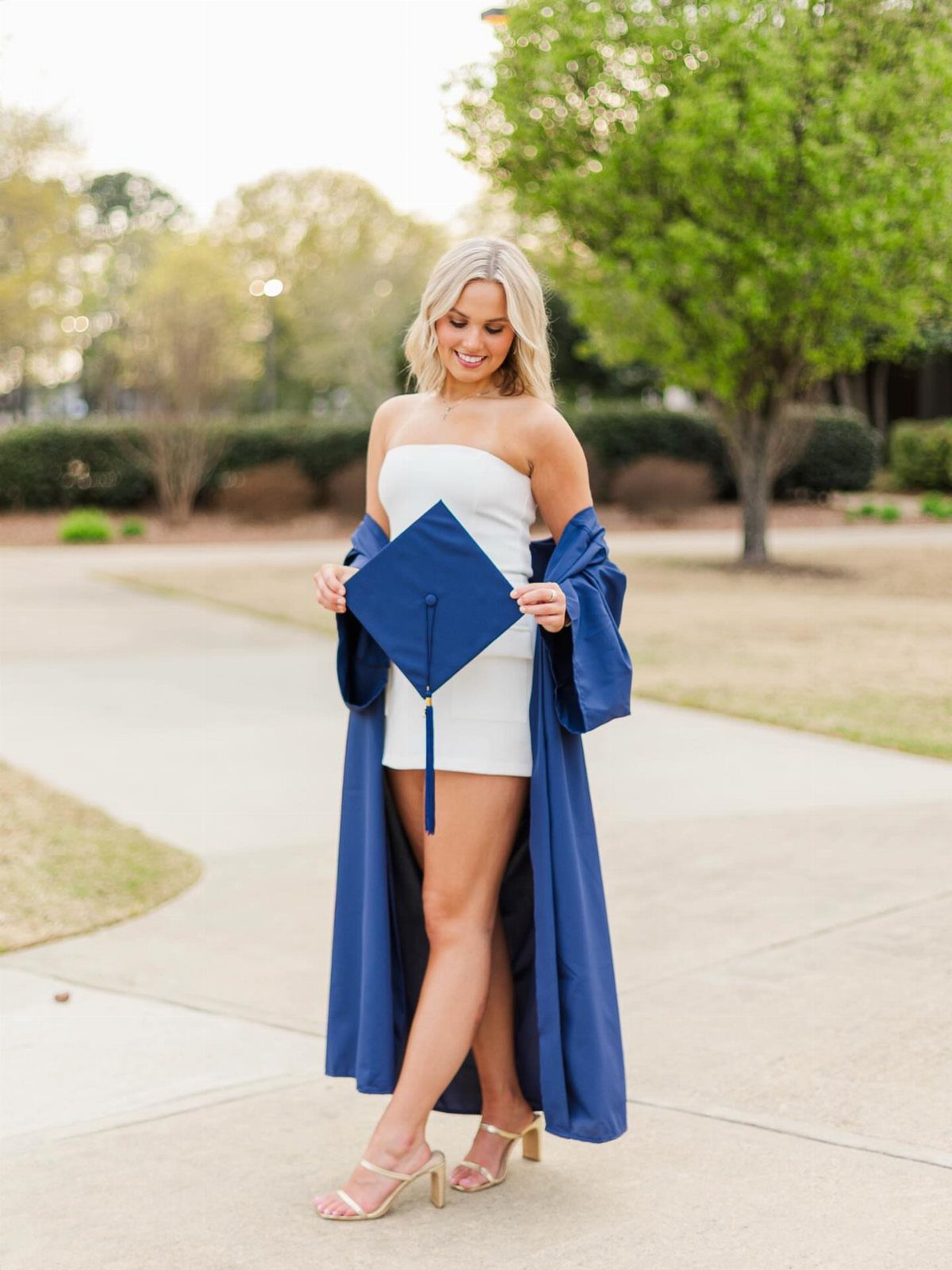 Woman in white dress with open blue graduation gown holding a cap outdoors