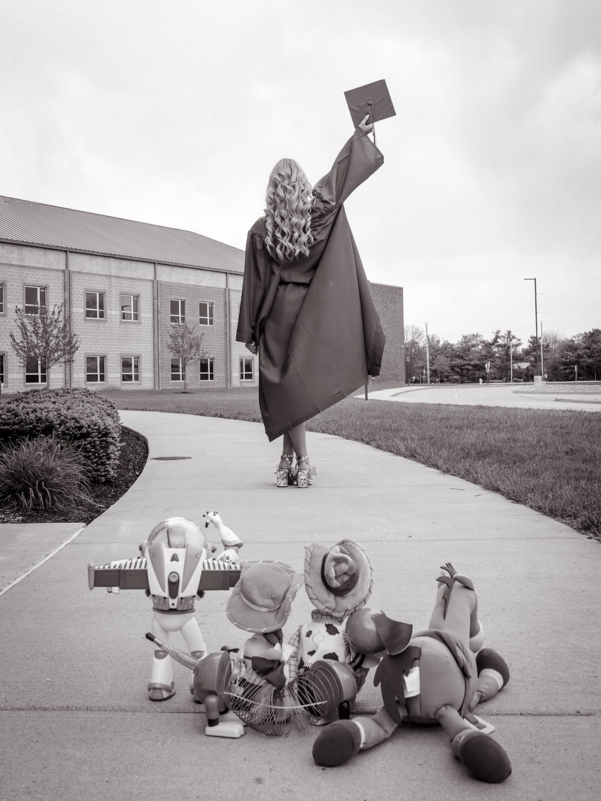 Woman in graduation cap and gown walking away on a sidewalk holding cap high