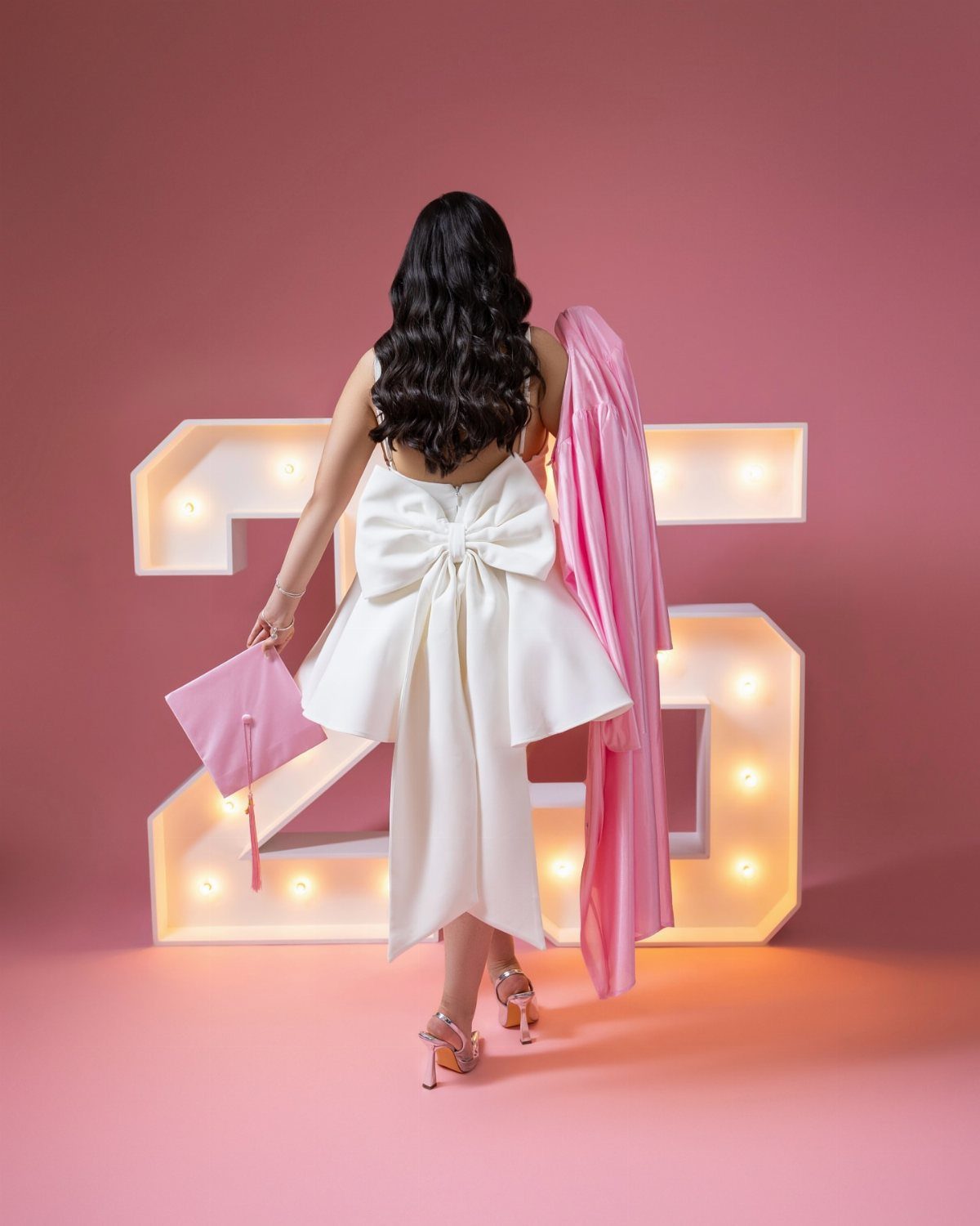 Woman in white midi dress with large bow holding pink graduation accessories.