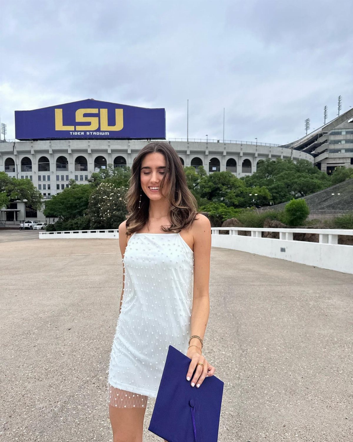 Young woman in a white dress holding a purple graduation cap before a stadium