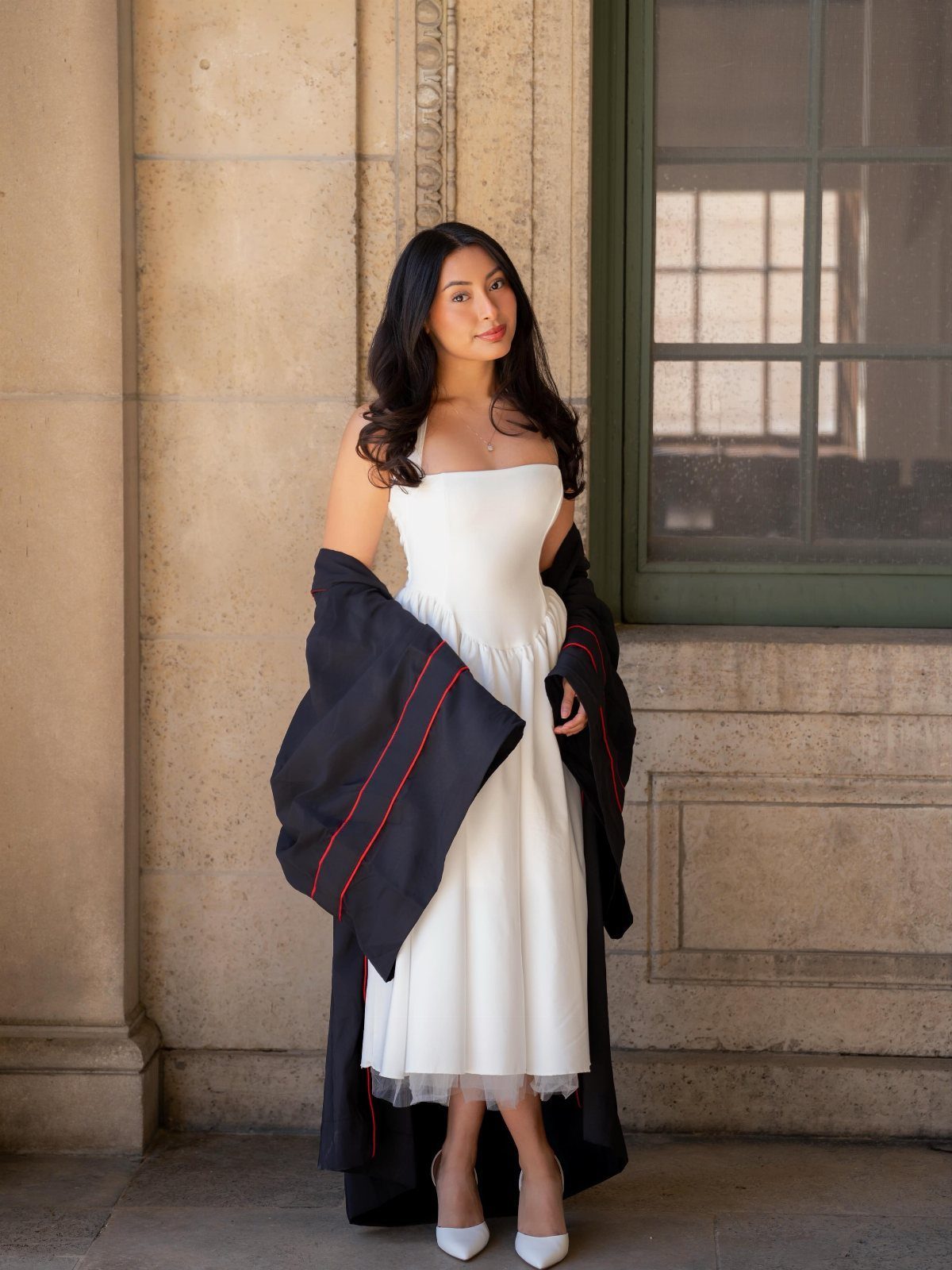 Young woman in a white midi dress and graduation gown posing by stone wall