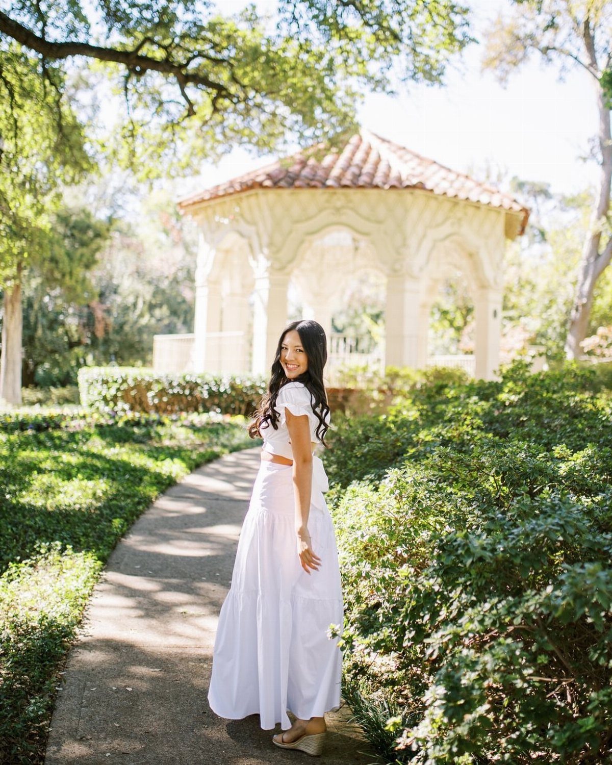 Young woman in a long white dress posing on a garden path near a gazebo