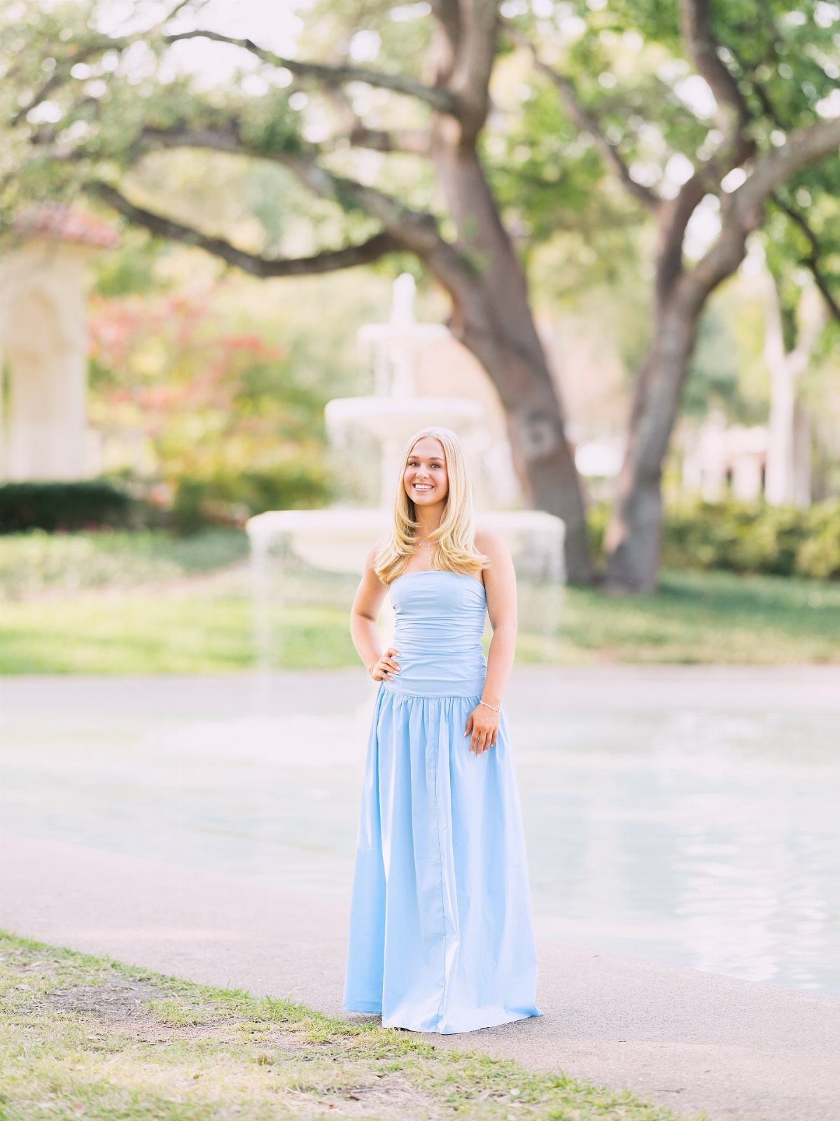 Young woman in a long light blue strapless dress posing by a park fountain