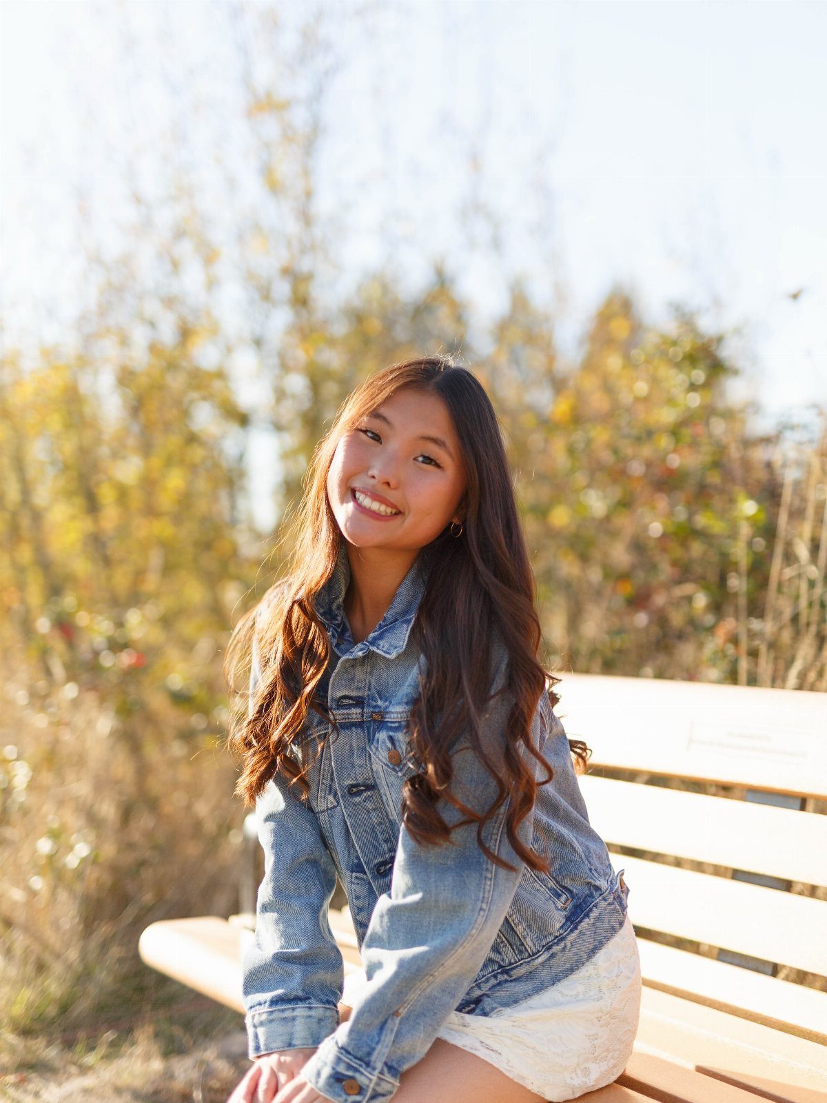 Young woman in a white lace dress and denim jacket sitting on a bench.