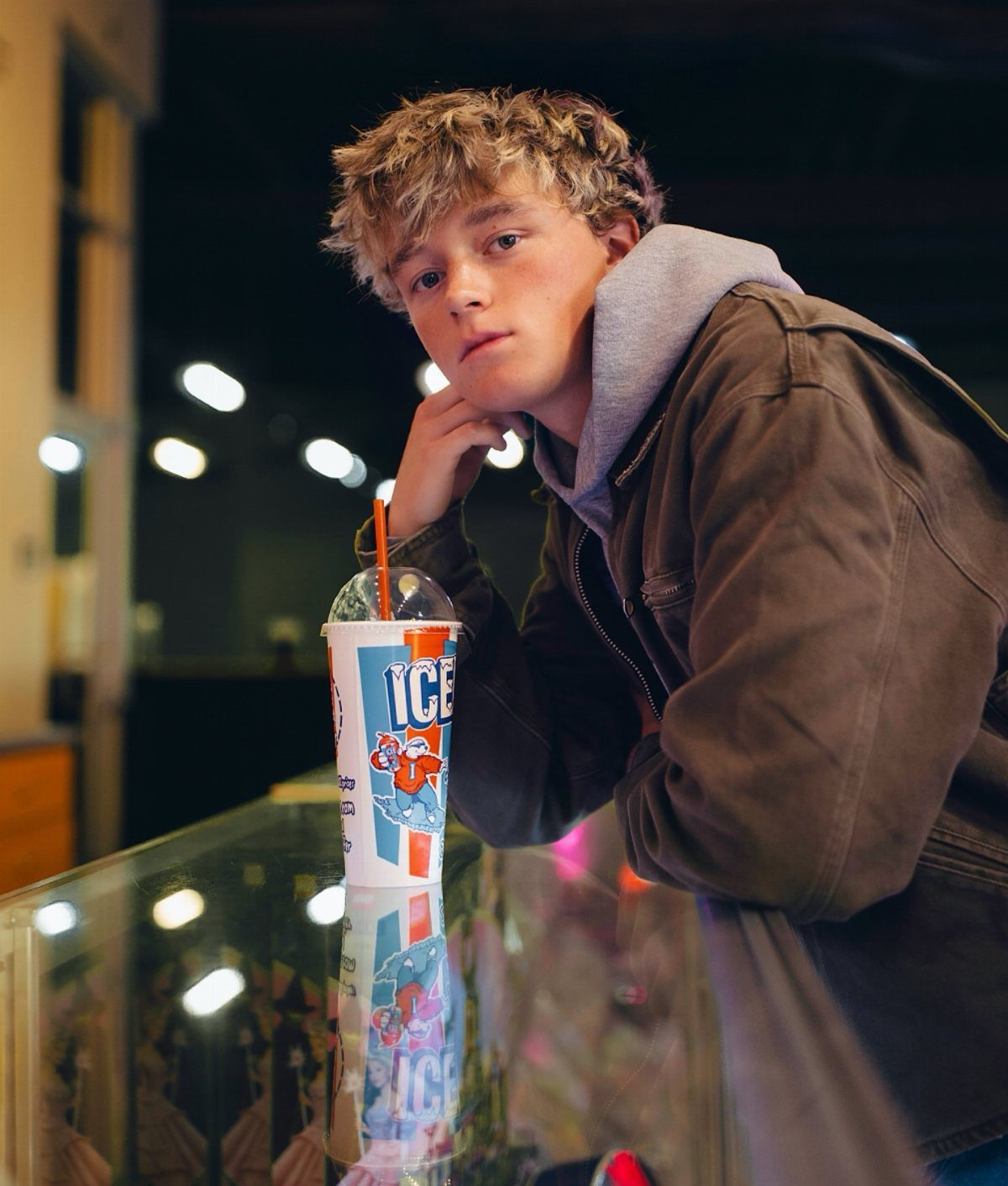 Teenage boy with curly blonde hair leaning on a counter with an ICEE drink.