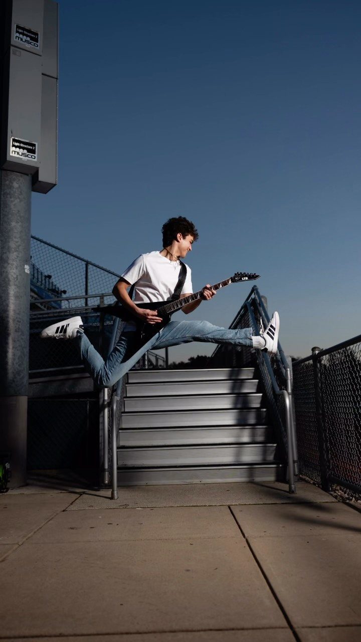 Young man jumping in the air while playing a black electric guitar outdoors.