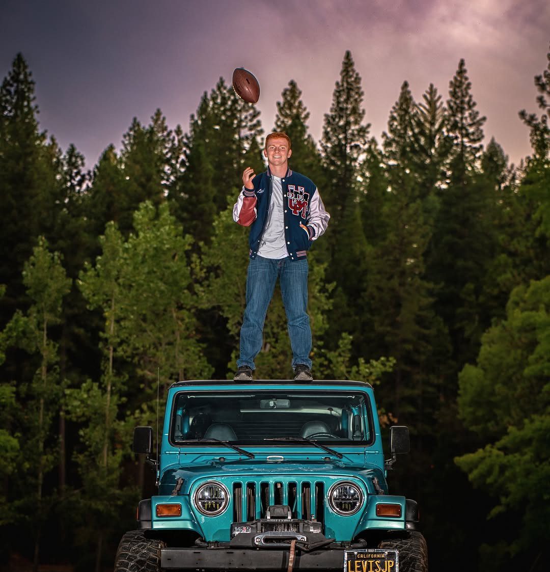 Young man in varsity jacket tossing football while standing on blue Jeep roof