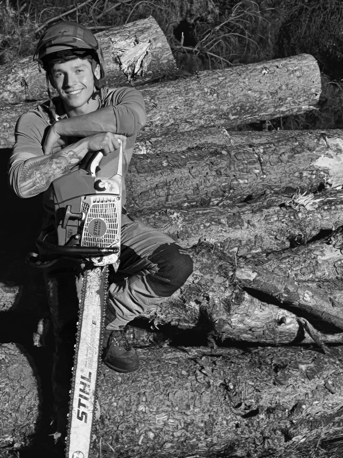 Smiling young man in a helmet leaning on a chainsaw among fallen logs.
