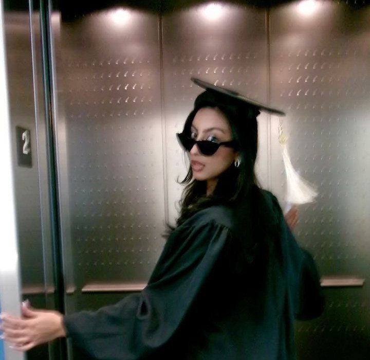 Young woman in a black graduation cap and gown posing in a metallic elevator