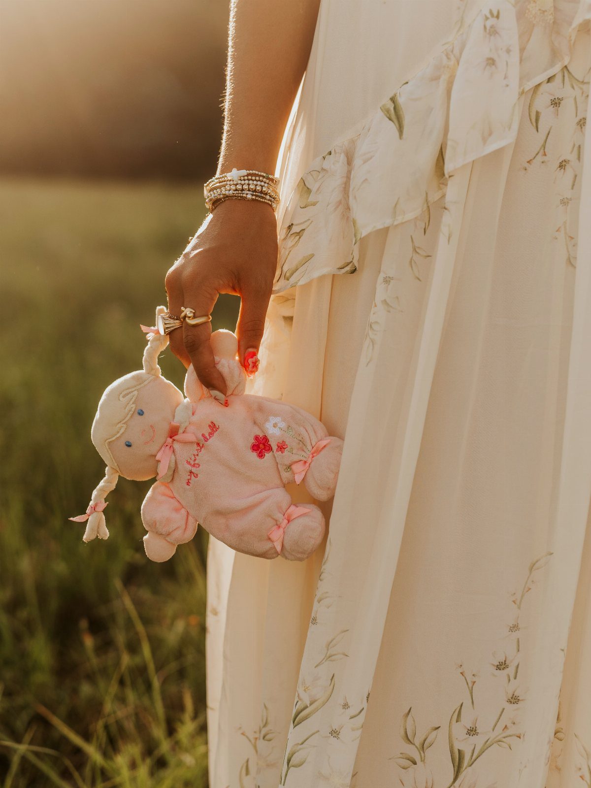 Hand holding a pink plush doll while wearing a floral cream dress outdoors.