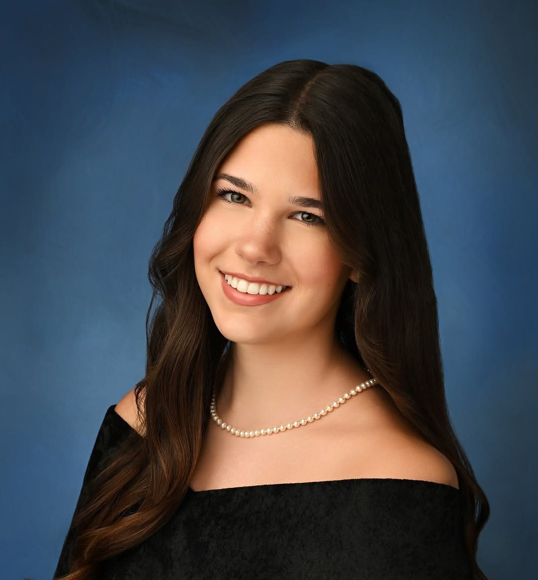 Smiling young woman with long dark hair wearing a pearl necklace and black top.