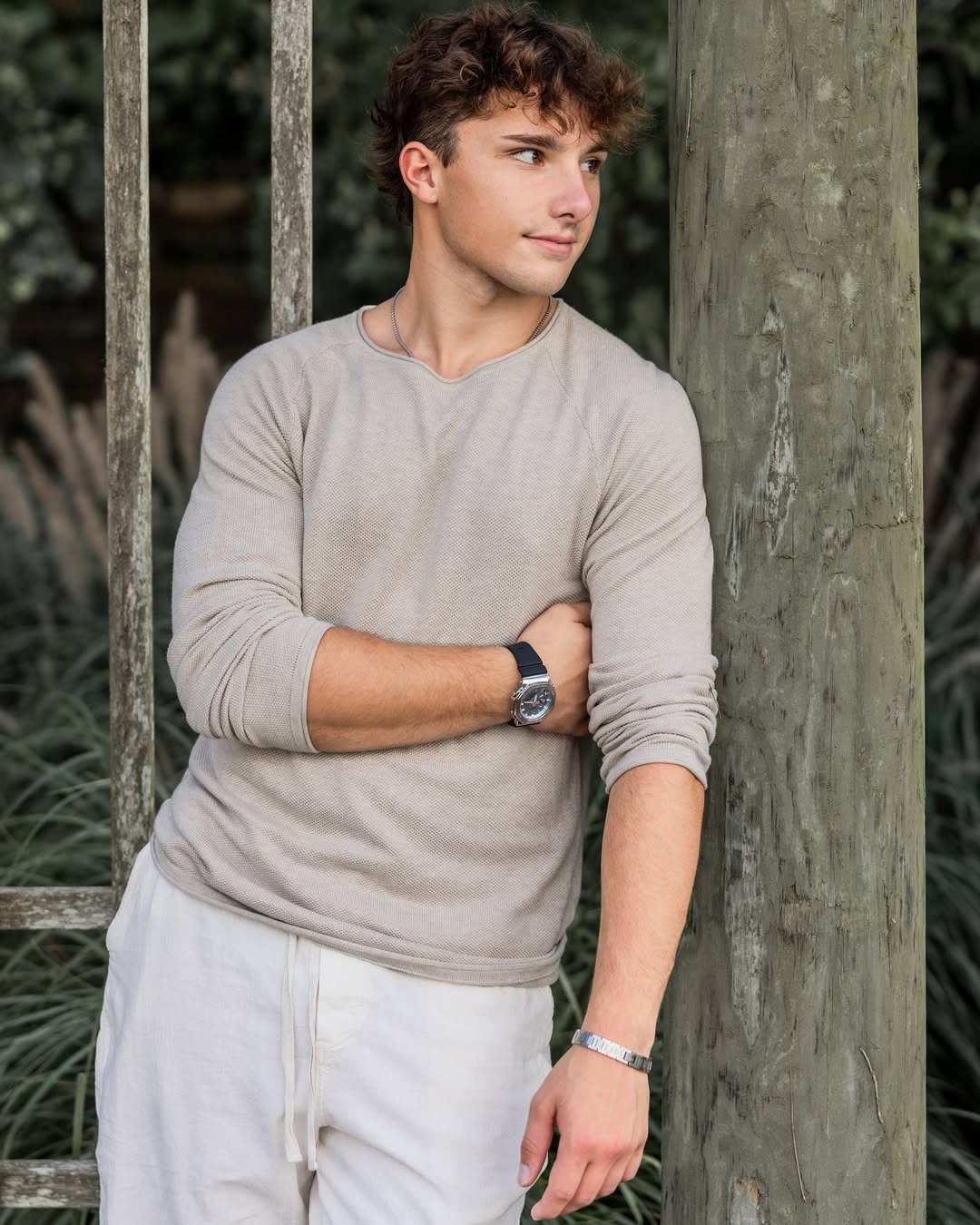 Young man in beige shirt and white pants leaning against a wooden post.