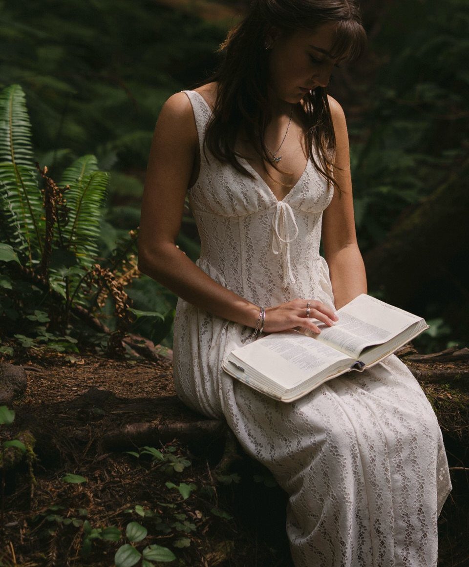 Young woman in white lace dress reading a book on a forest log