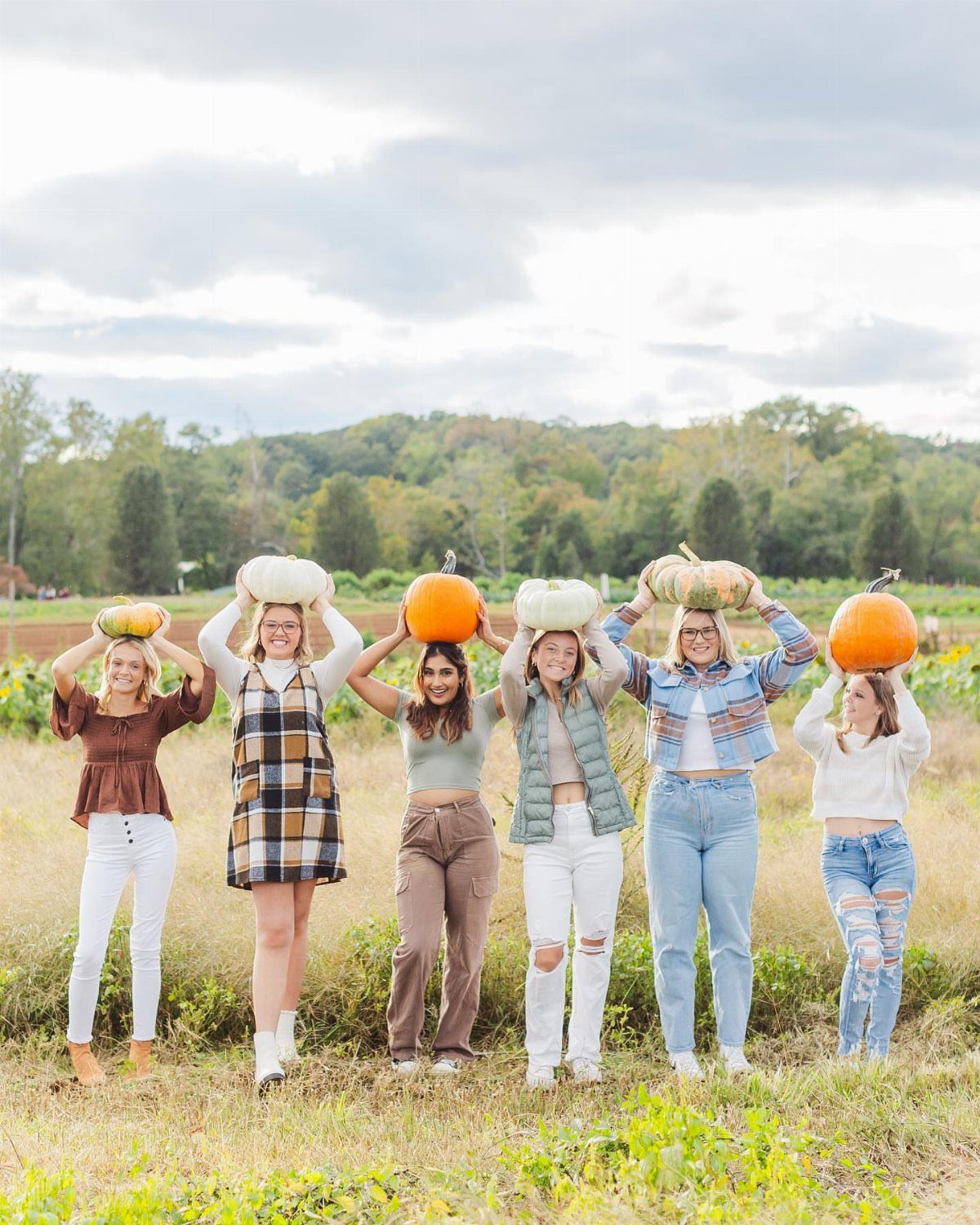 Six young women balancing pumpkins on their heads in a grassy autumn field.