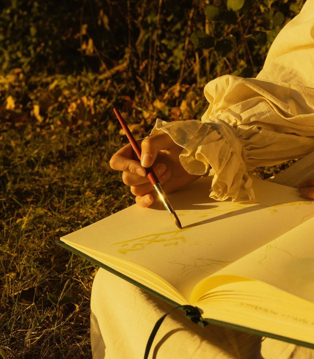 Person in white ruffled clothing painting in a sketchbook outdoors during golden hour