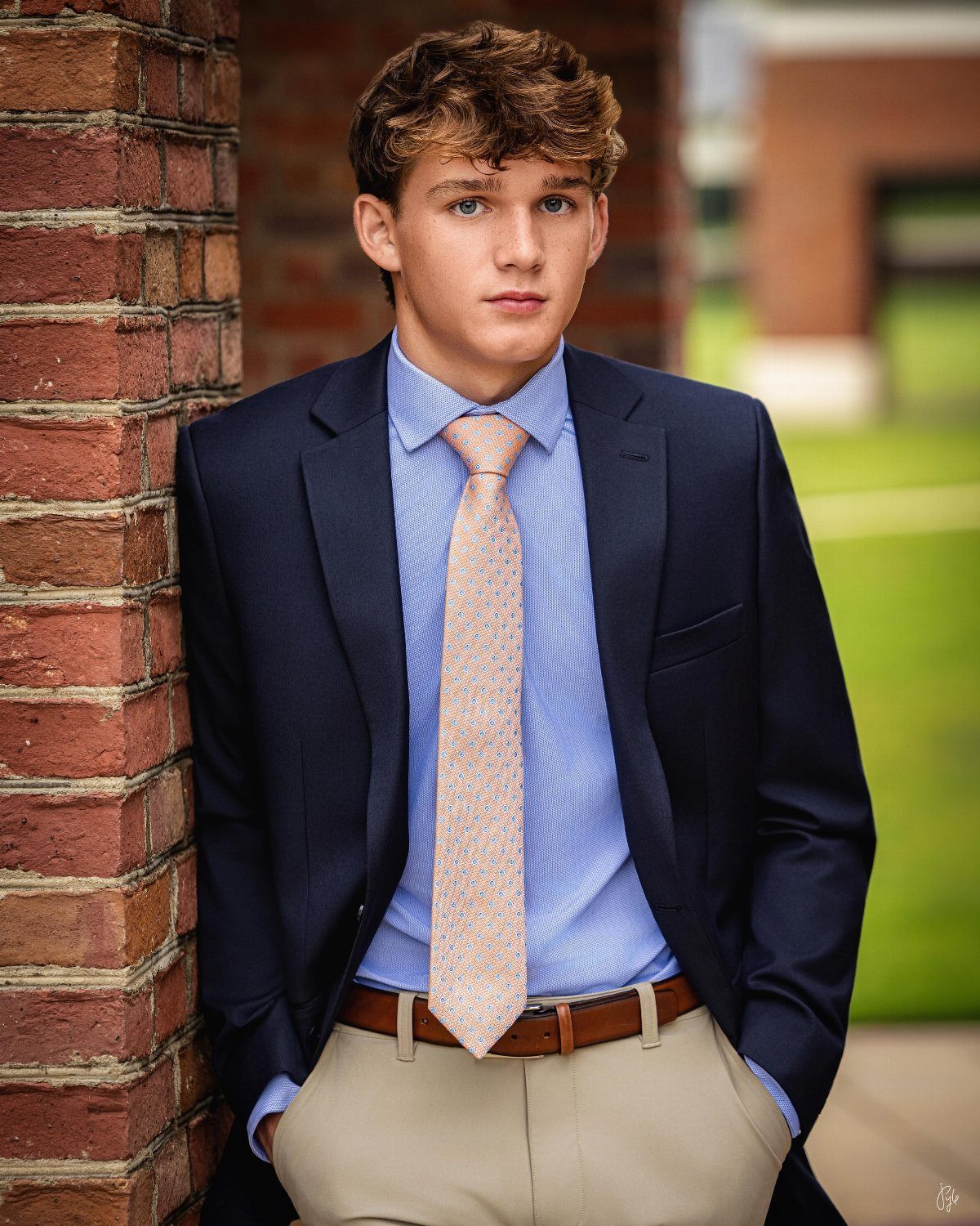 Young man in a navy blazer and orange tie leaning against brick wall