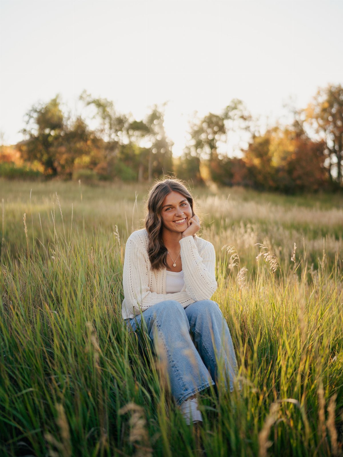 Young woman smiling in a grassy field with autumn foliage in the background.