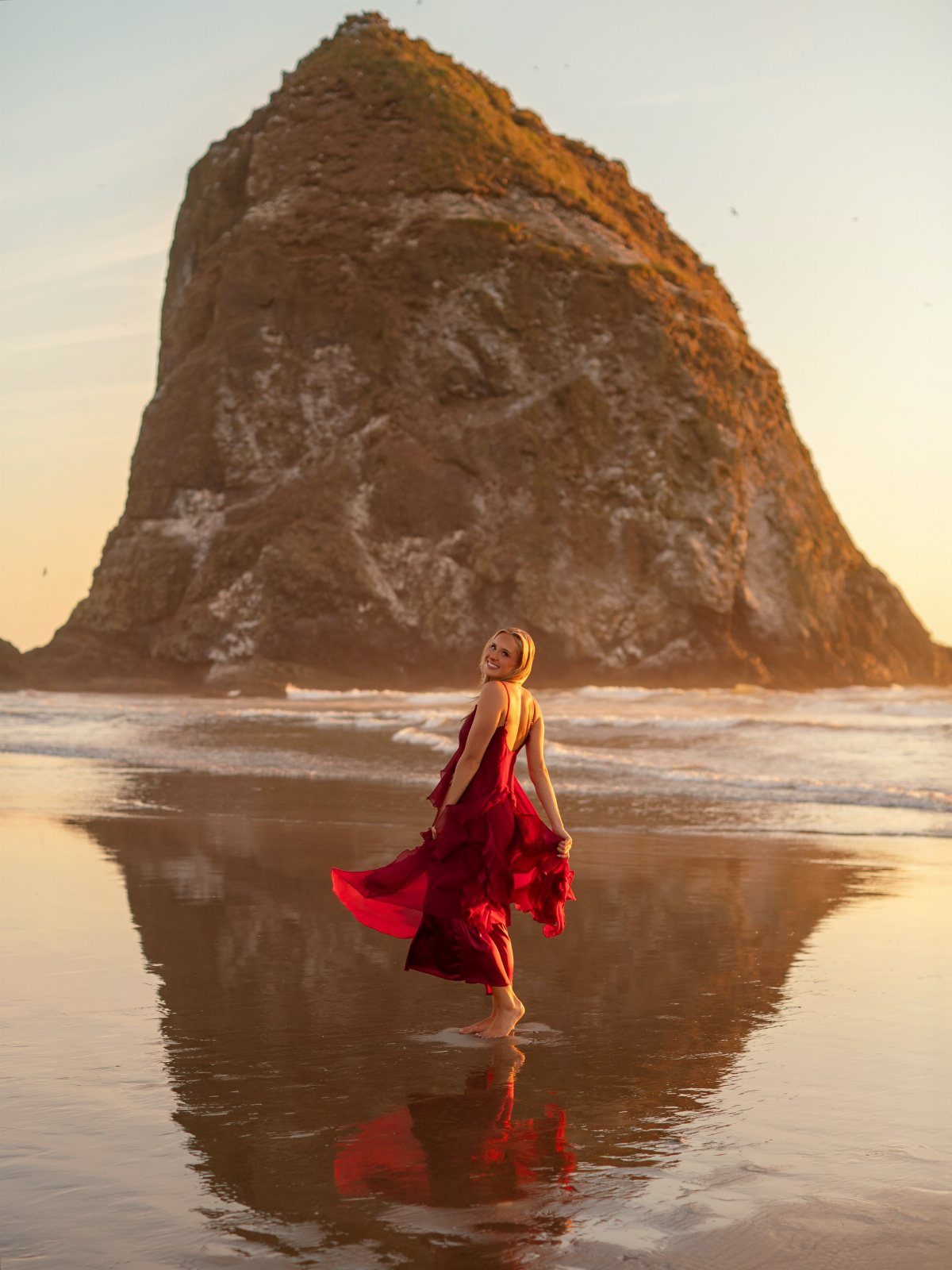 Woman in a flowing red dress standing on a beach before a large rock