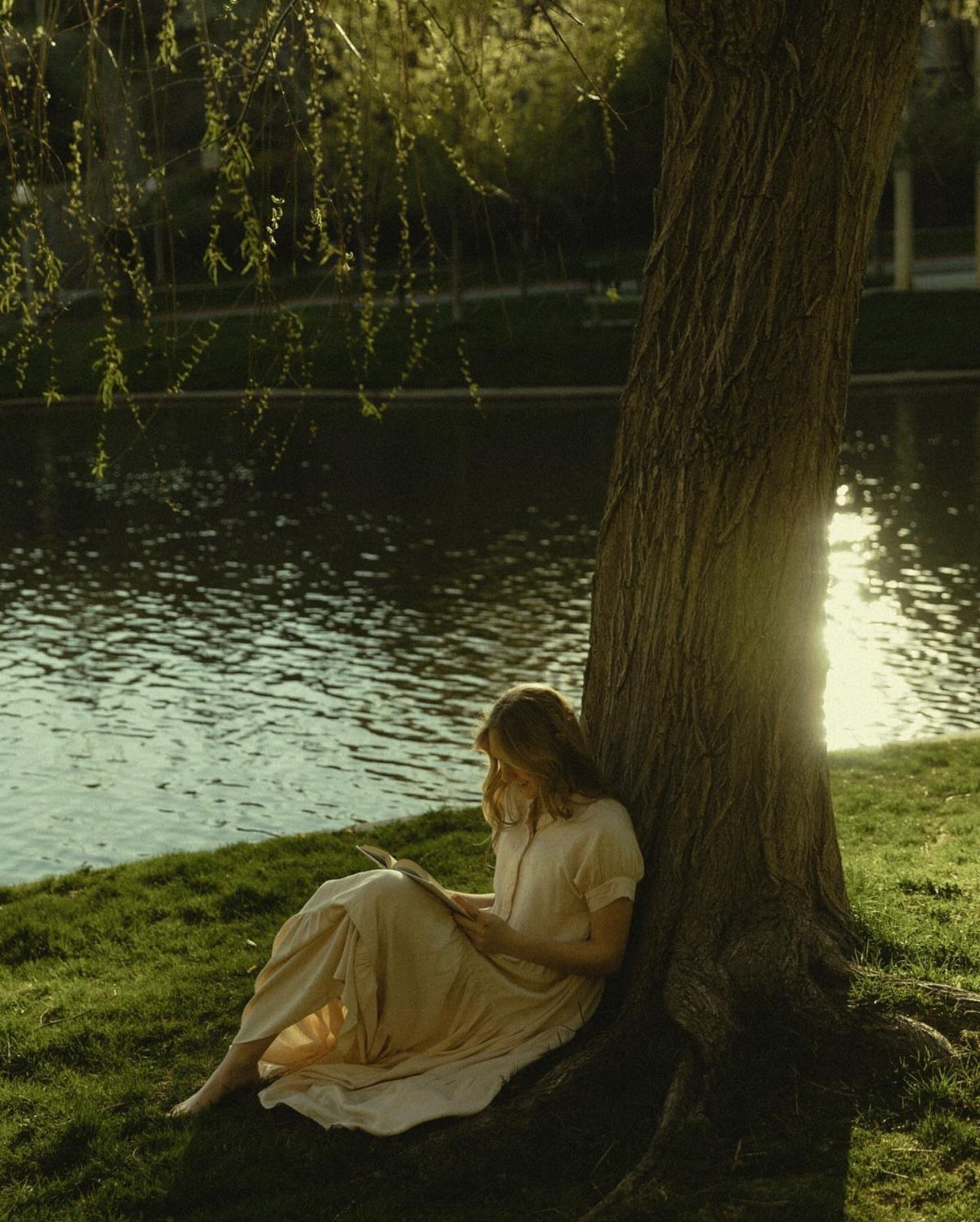 Woman in cream dress reading a book while leaning against a tree by water