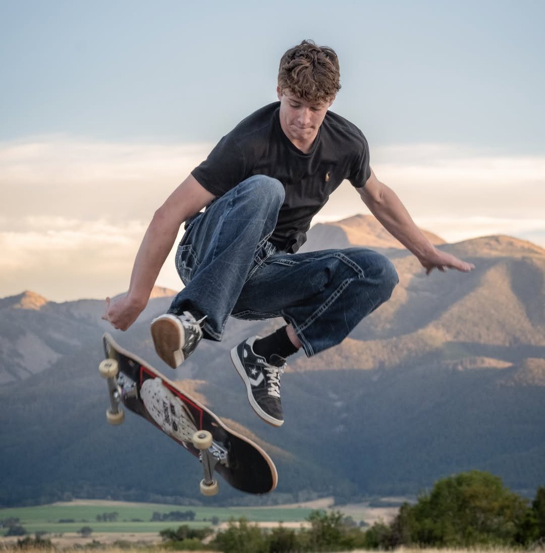 Young man jumping mid-air on a skateboard with mountains in the background.