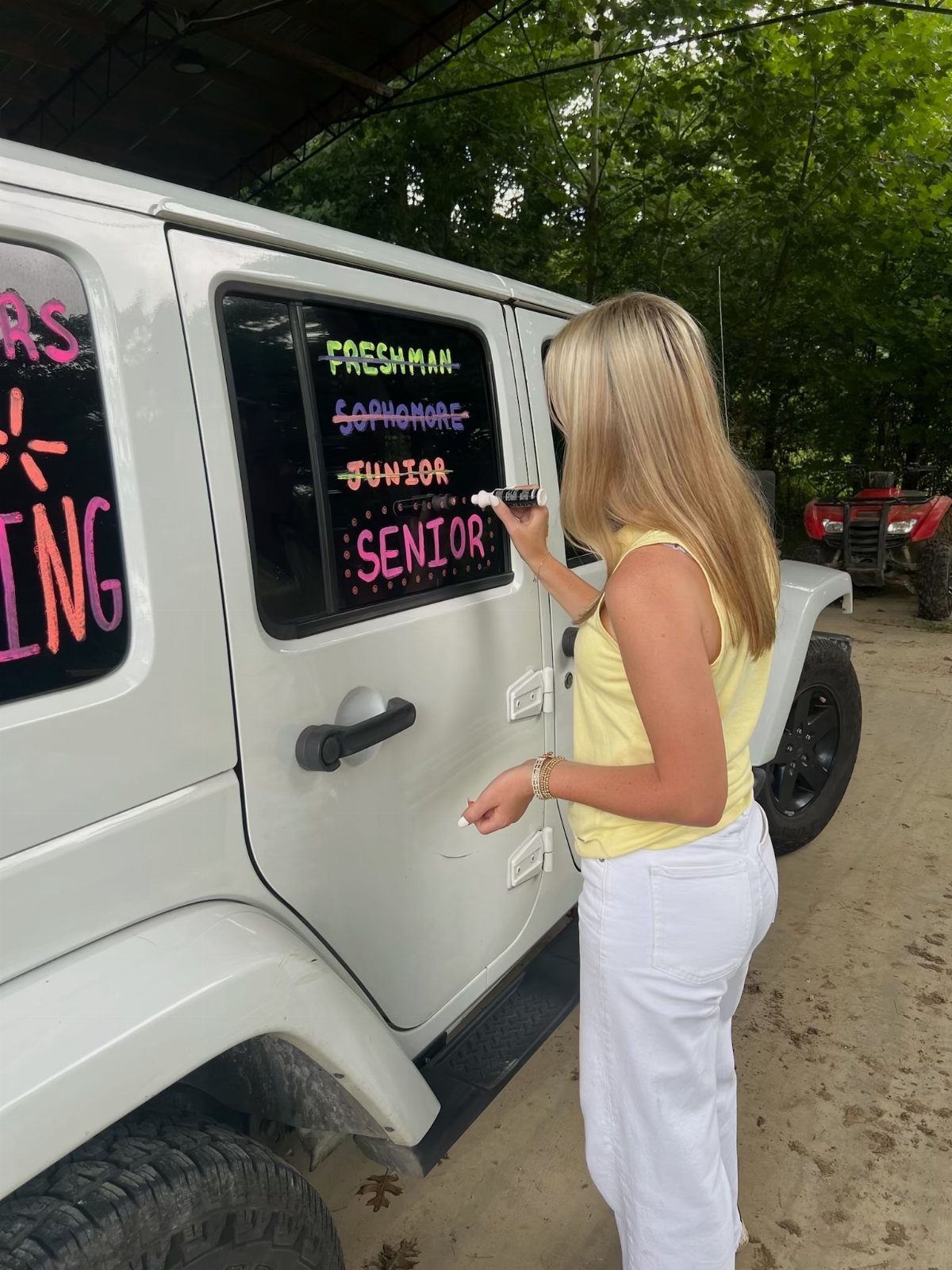 Woman writing senior year milestones on the window of a white Jeep