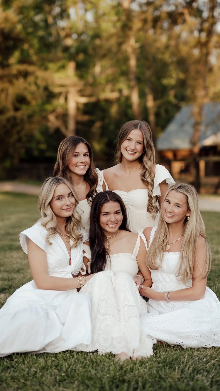 Five young women in white dresses smiling and posing together on a grassy lawn.