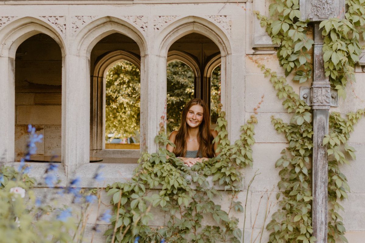Young woman smiling from a stone arched window surrounded by green climbing vines.