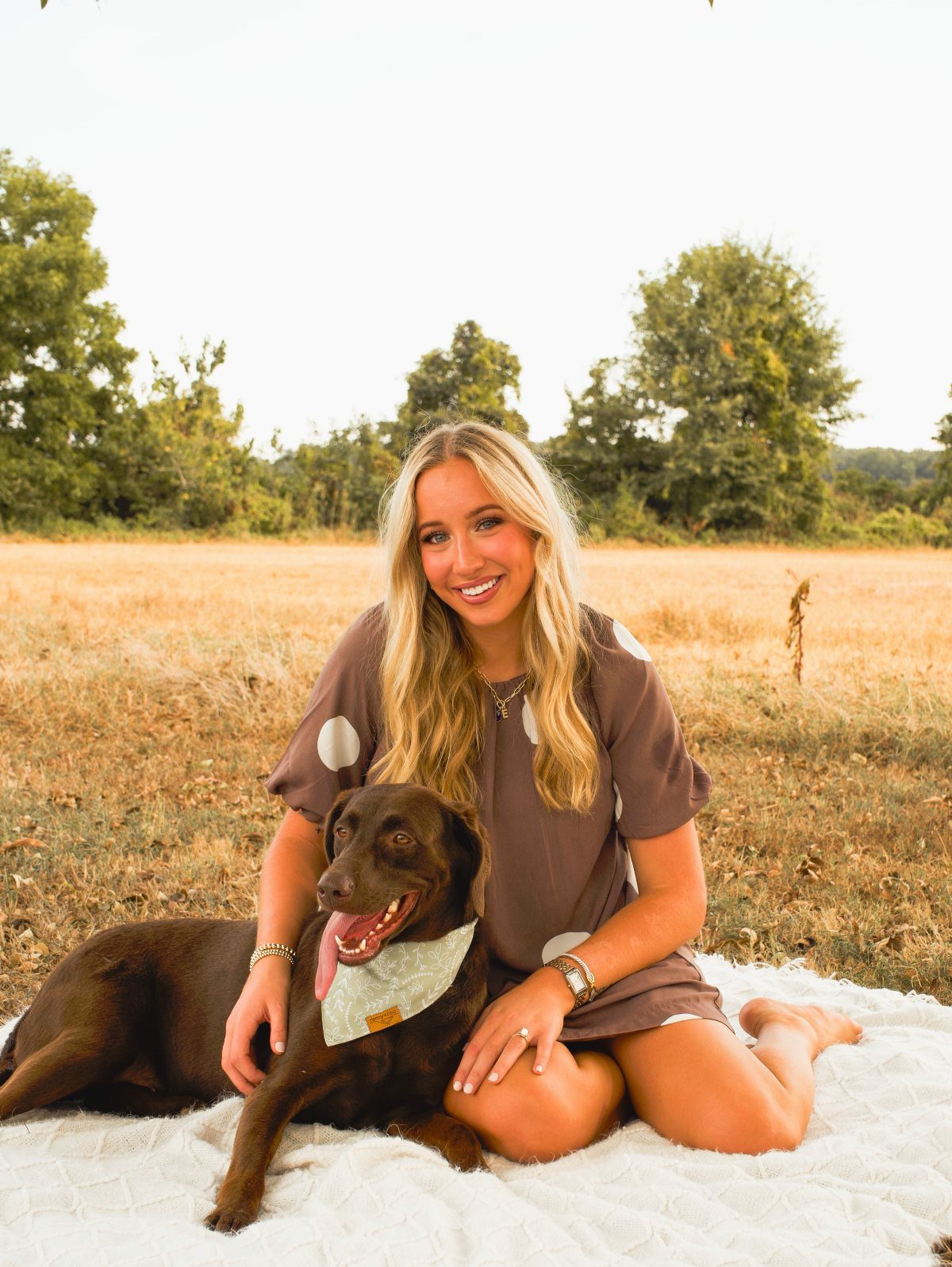 Young woman in a brown dress sitting with a chocolate Labrador in a field