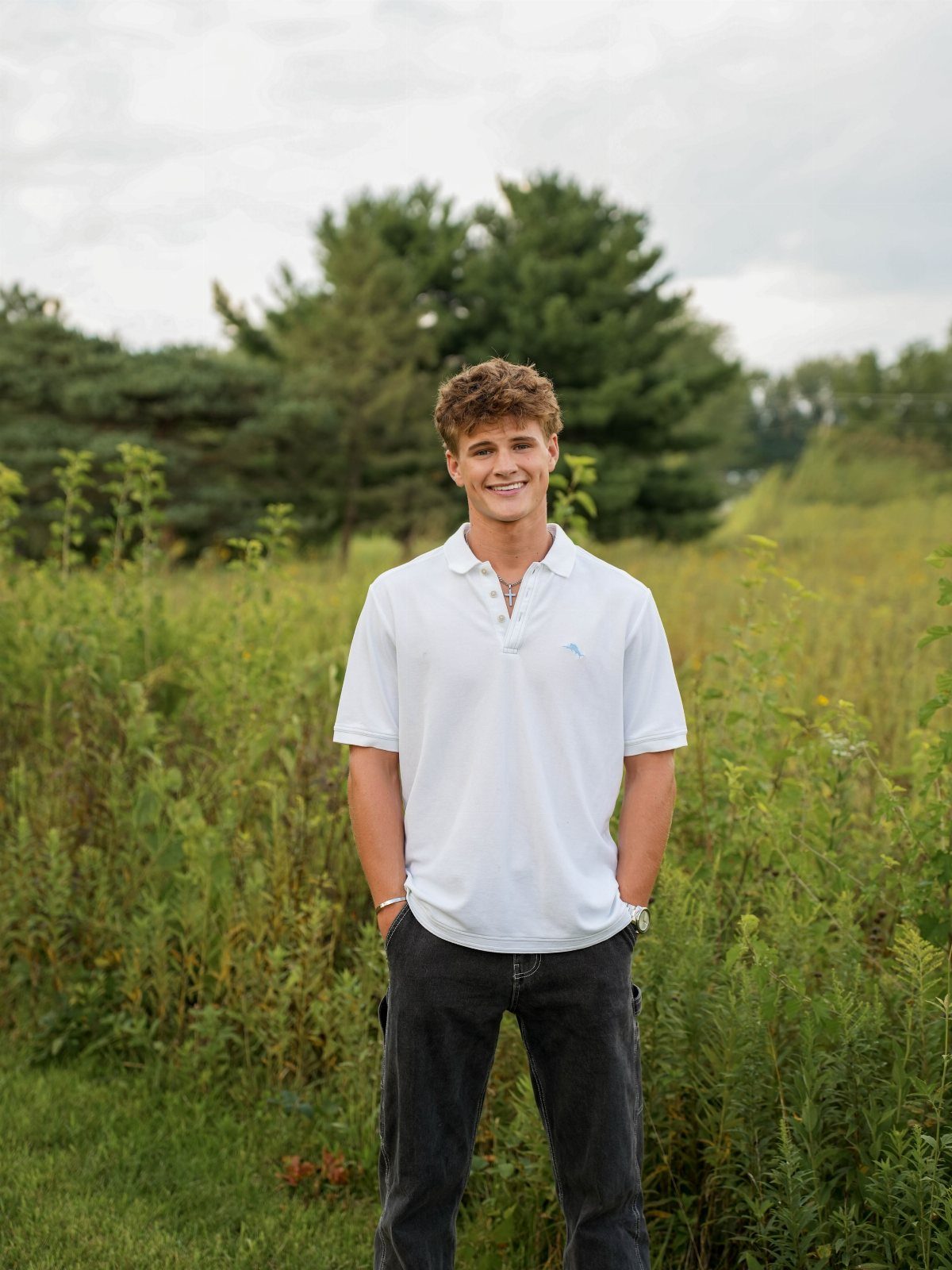 Young man in white polo shirt posing in a grassy field with trees.