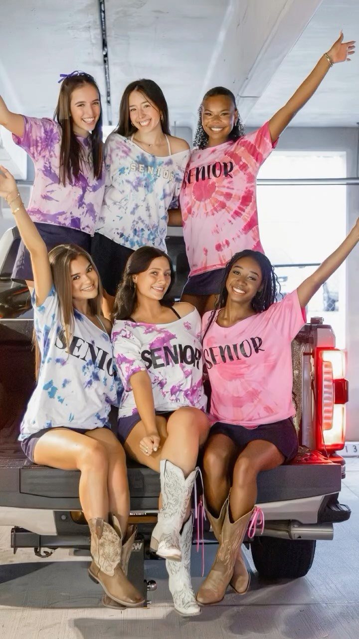 Six teenage girls in senior tie-dye shirts posing on a truck in a garage