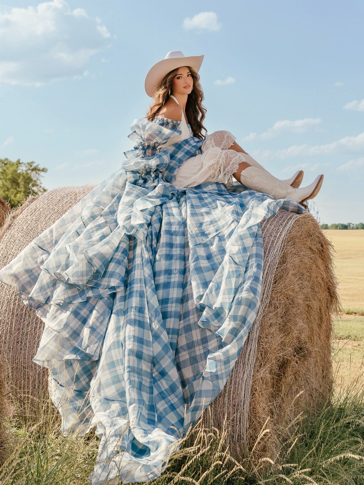 Woman in a blue gingham gown and cowboy hat sitting on a hay bale.