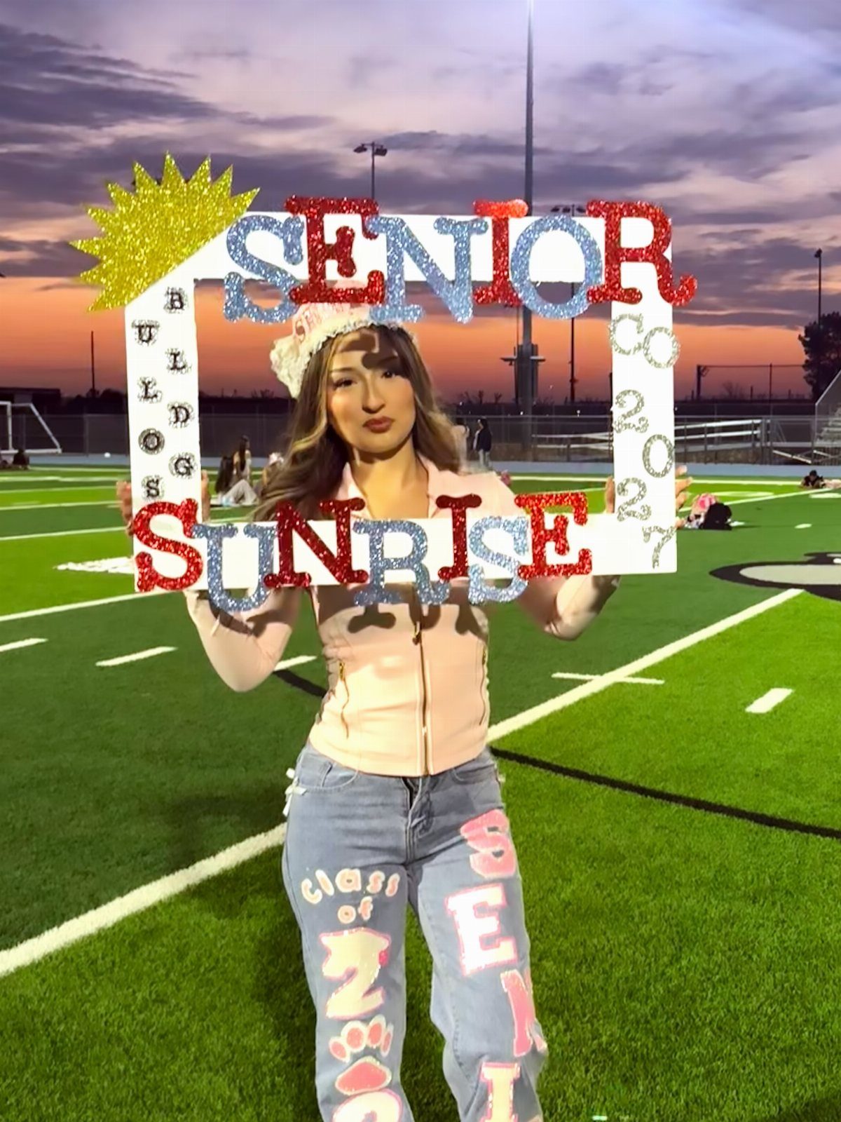 Young woman holding a Senior Sunrise frame on a green sports field at dawn