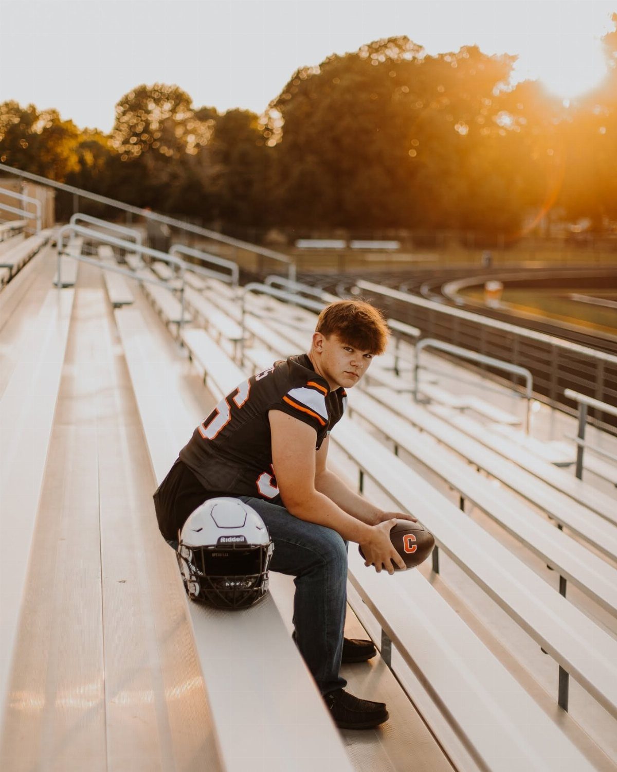 Teenage football player sitting on stadium bleachers holding a football during golden hour.