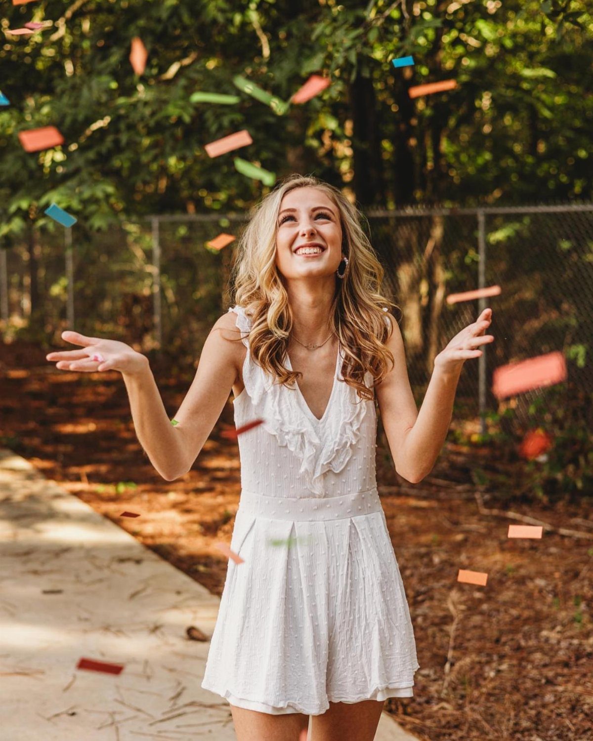 Smiling young woman in a white dress surrounded by floating colorful confetti outdoors.