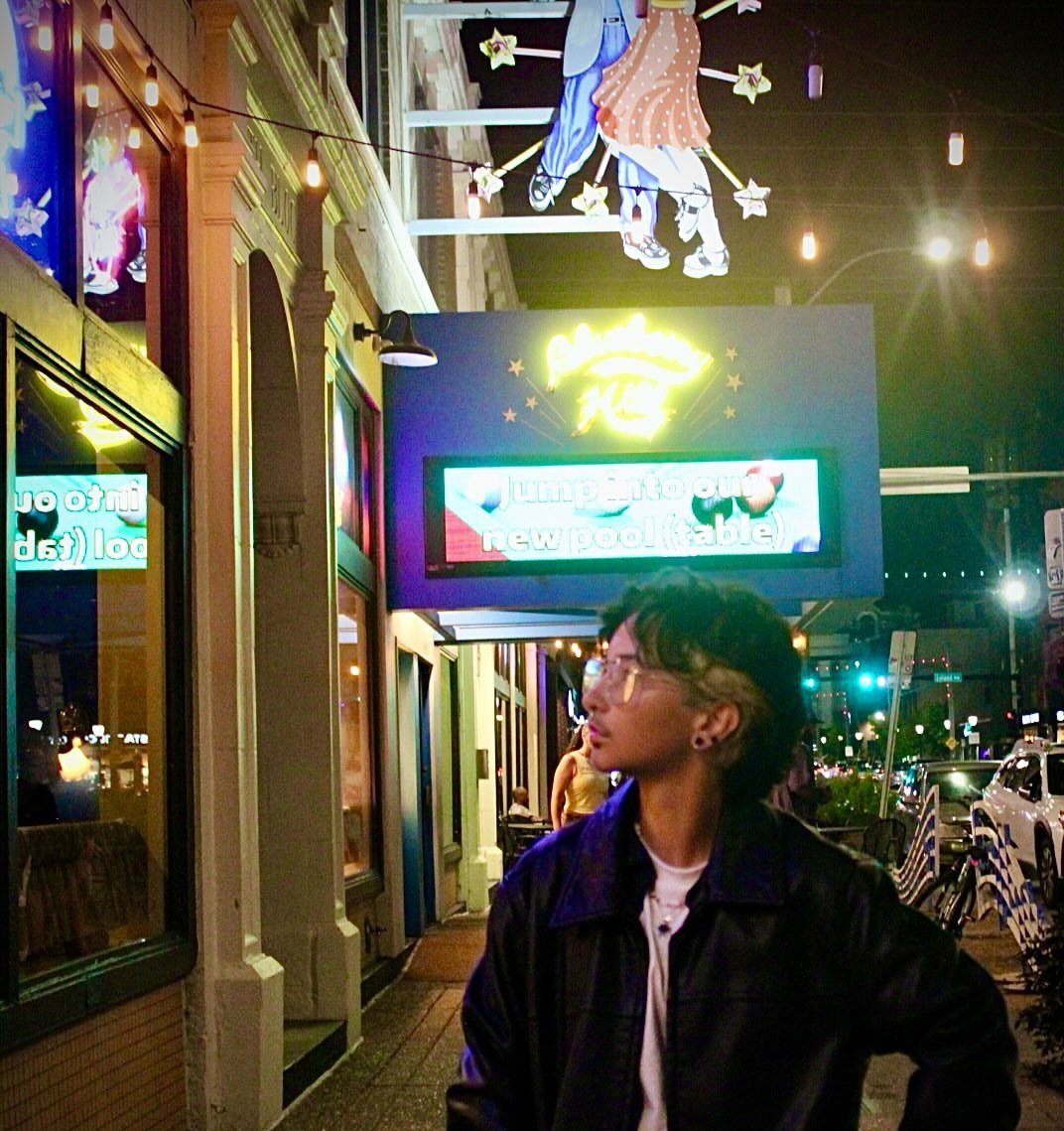 Young person in leather jacket posing on a city street with neon signs.