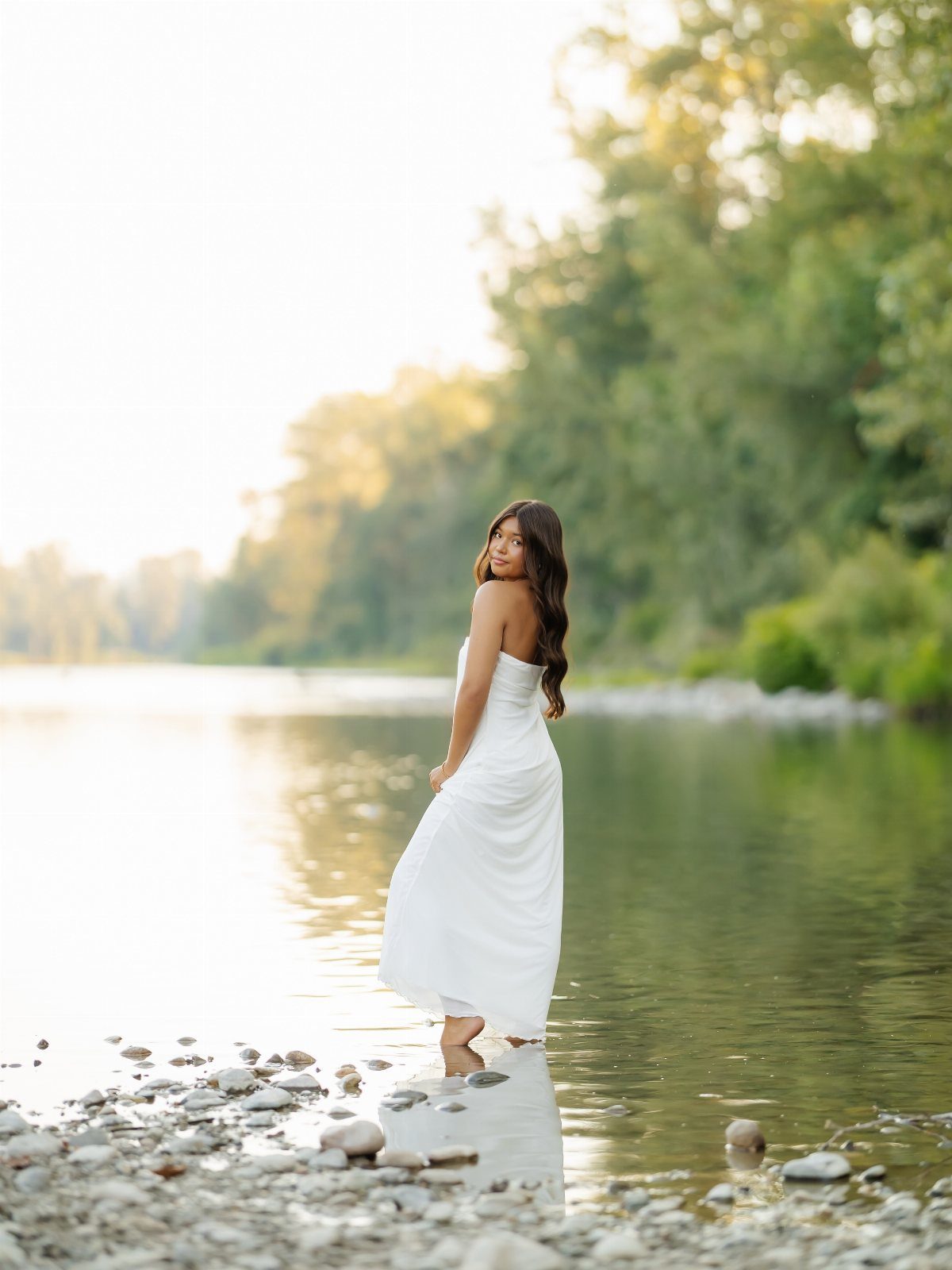 Young woman in a white dress standing in a shallow river by green trees