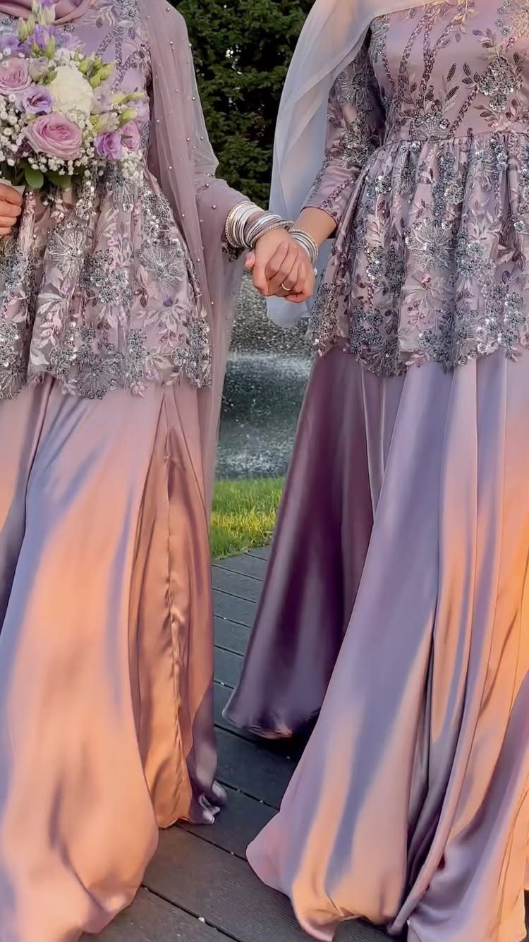 Two people in matching lavender embroidered gowns holding hands outdoors with a bouquet.