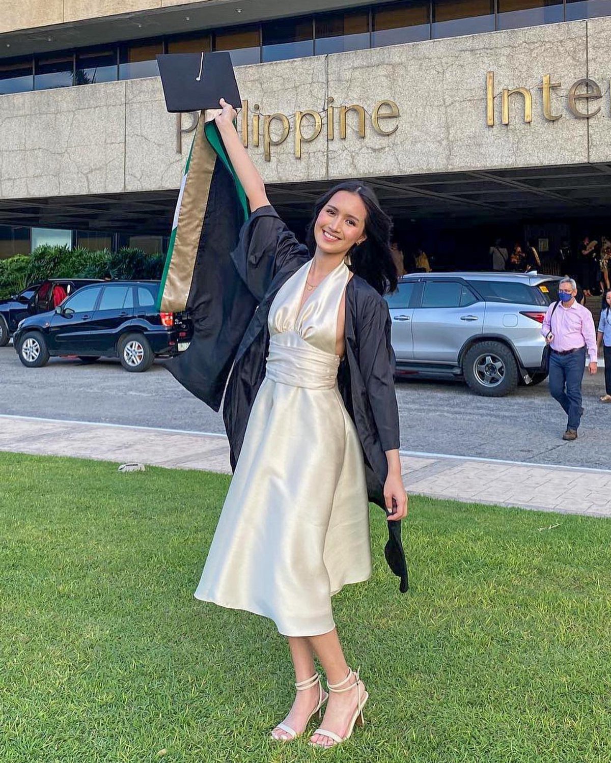 Woman in a cream satin midi dress holding a graduation cap on a lawn